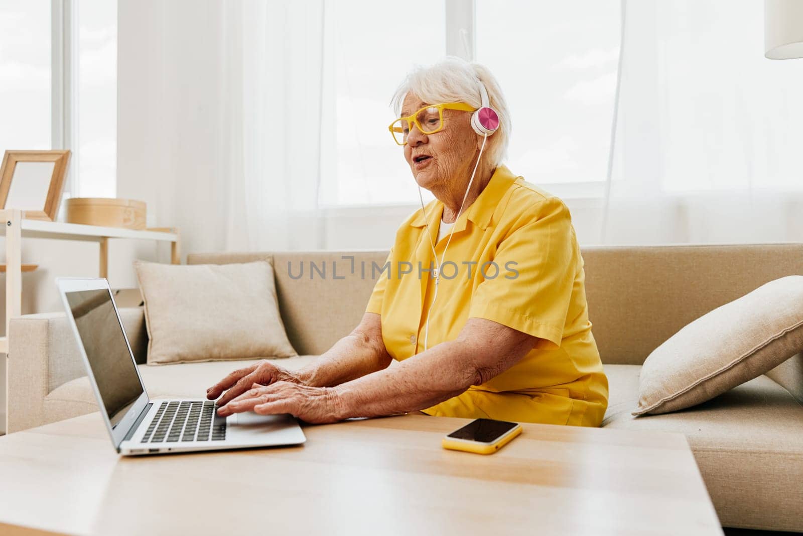 Happy elderly woman with a laptop typing in headphones smile sitting at home on the couch in a yellow shirt, bright modern interior, lifestyle online communication. High quality photo