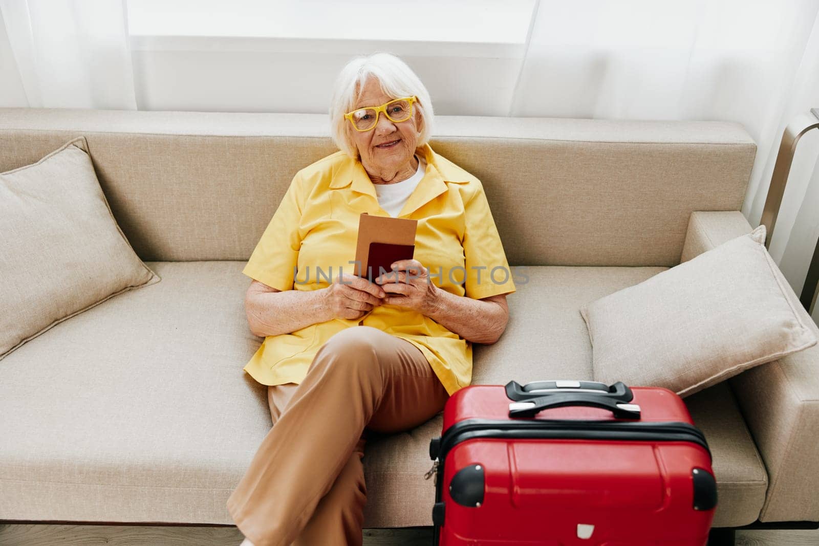 Happy senior woman with passport and travel ticket packed a red suitcase, vacation and health care. Smiling old woman happily sits on the sofa before the trip. High quality photo
