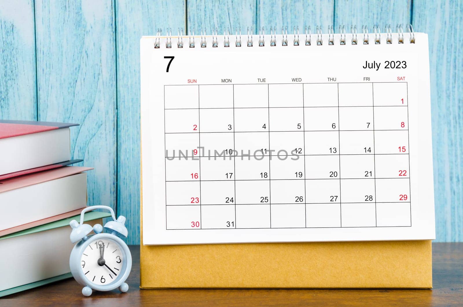 July 2023 Monthly desk calendar for the organizer to plan 2023 year with alarm clock and books on wooden table.