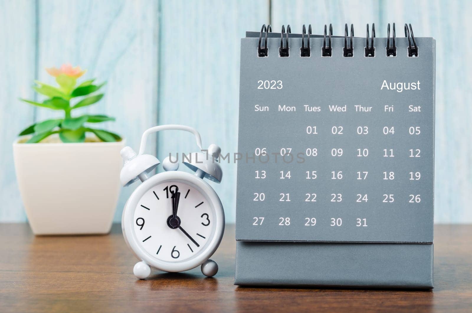 The August 2023 Monthly desk calendar for 2023 year with alarm clock on blue wooden background. by Gamjai