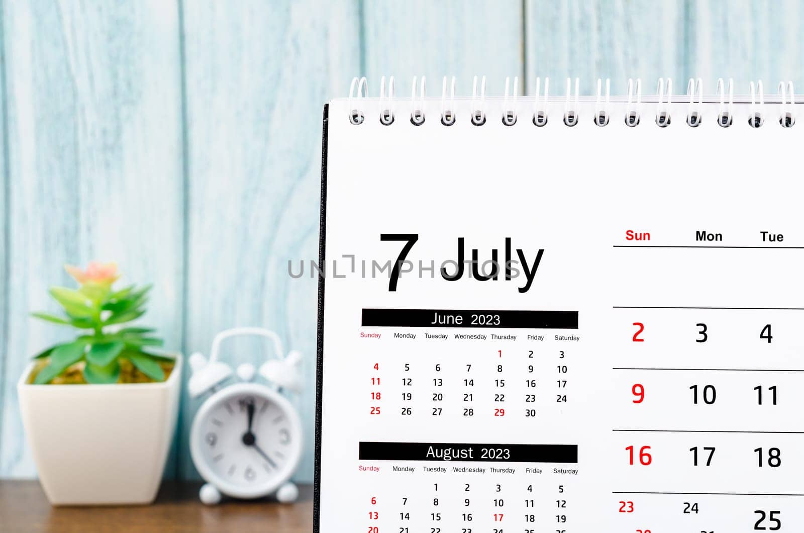 July 2023 Monthly desk calendar for 2023 year with alarm clock on blue wooden background.