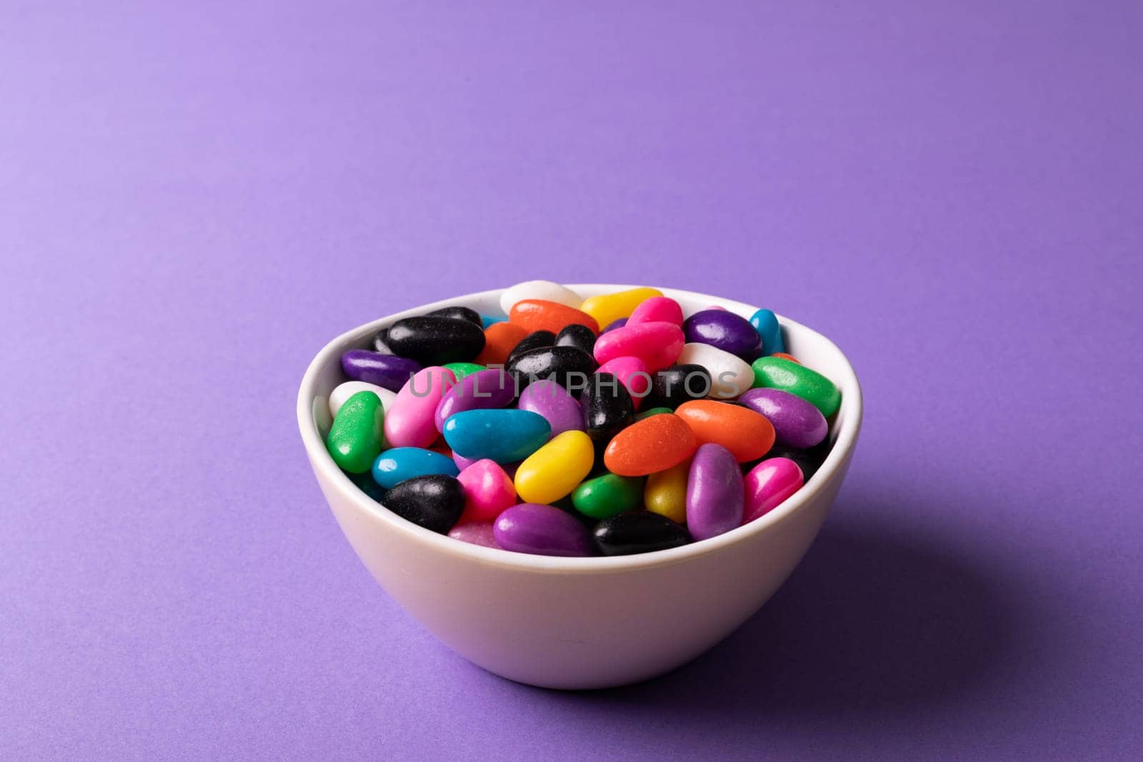Close-up of multi colored candies in white bowl on purple background with copy space. unaltered, sweet food and unhealthy eating concept.
