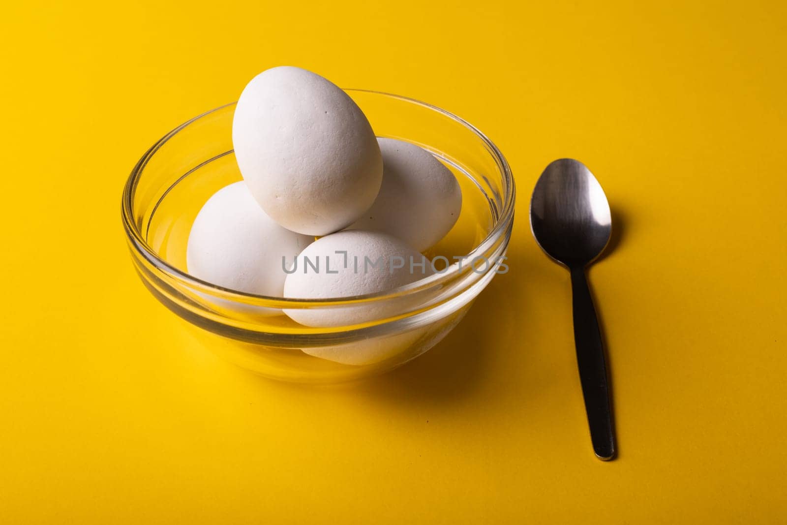 High angle close-up of fresh white eggs in glass bowl by spoon on yellow background. unaltered, food, healthy eating concept.