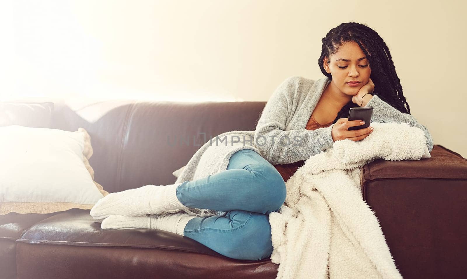 Patiently waiting on him to text back. a young woman using her phone while relaxing in her her living room. by YuriArcurs