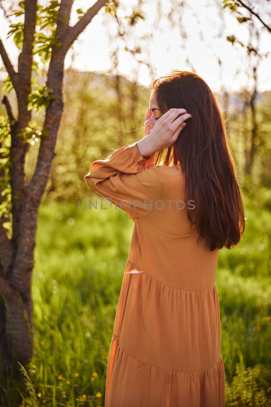 dear, a woman is standing in nature near a tree in a long orange dress, illuminated from behind by the sunset rays of the sun and straightens her hair behind her ear with her hand. Vertical photography. High quality photo