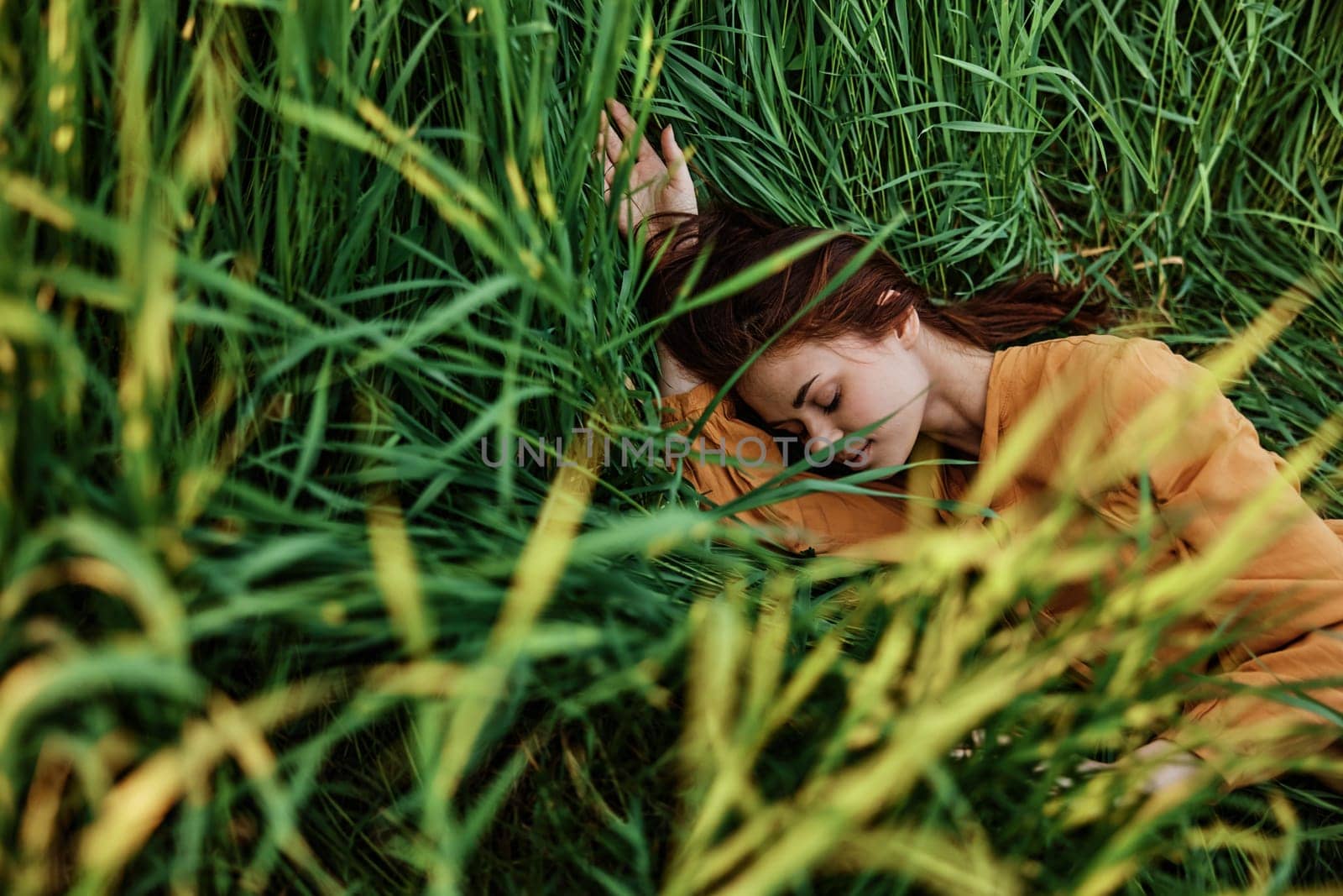 a pleasant woman in a long orange dress resting lying in the tall grass with her eyes closed in sunny weather at sunset with her arms outstretched. Street photography, the theme of privacy with nature by Vichizh