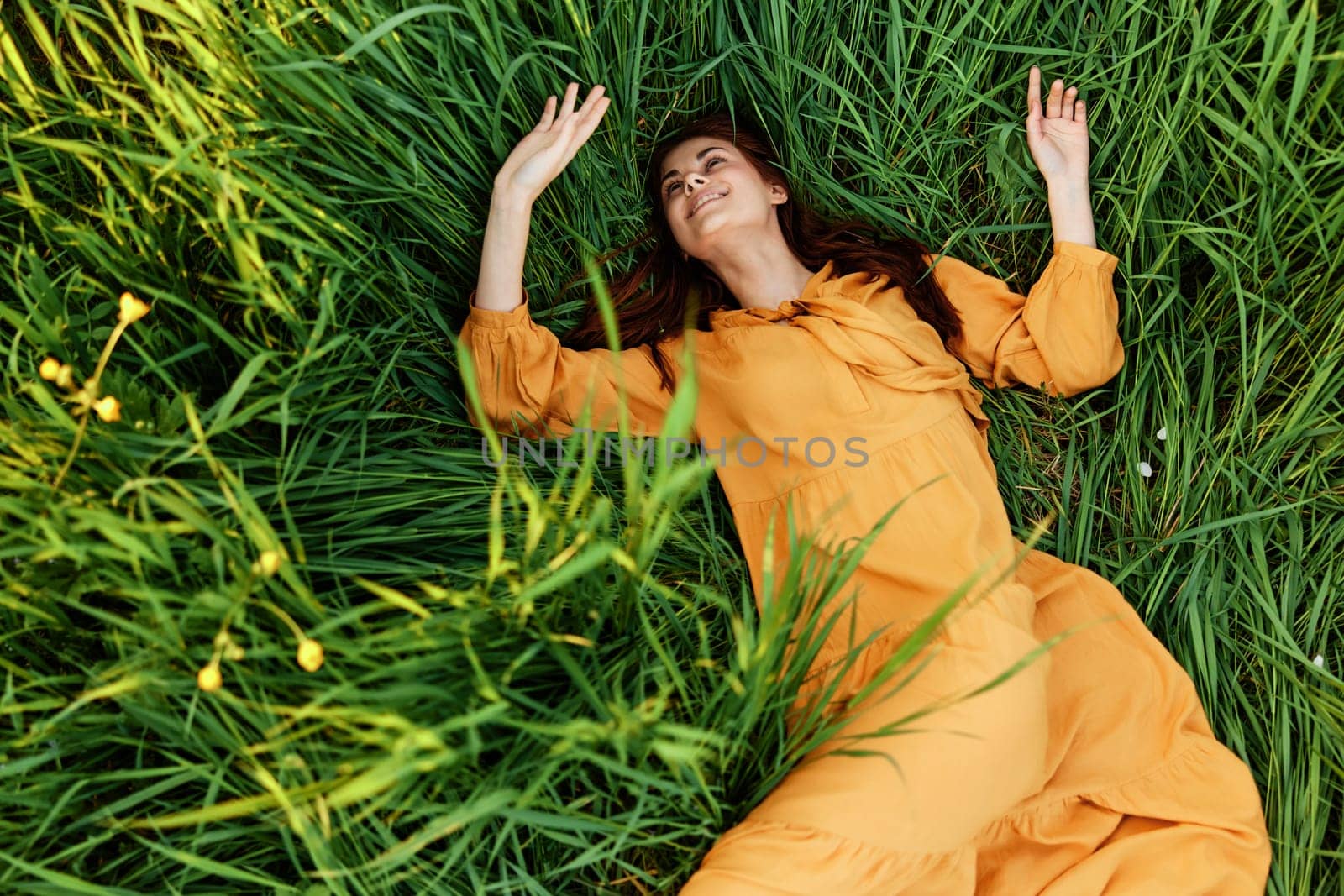 a relaxed red-haired woman enjoys summer lying in the tall green grass in a long orange dress smiling happily with her eyes closed by Vichizh