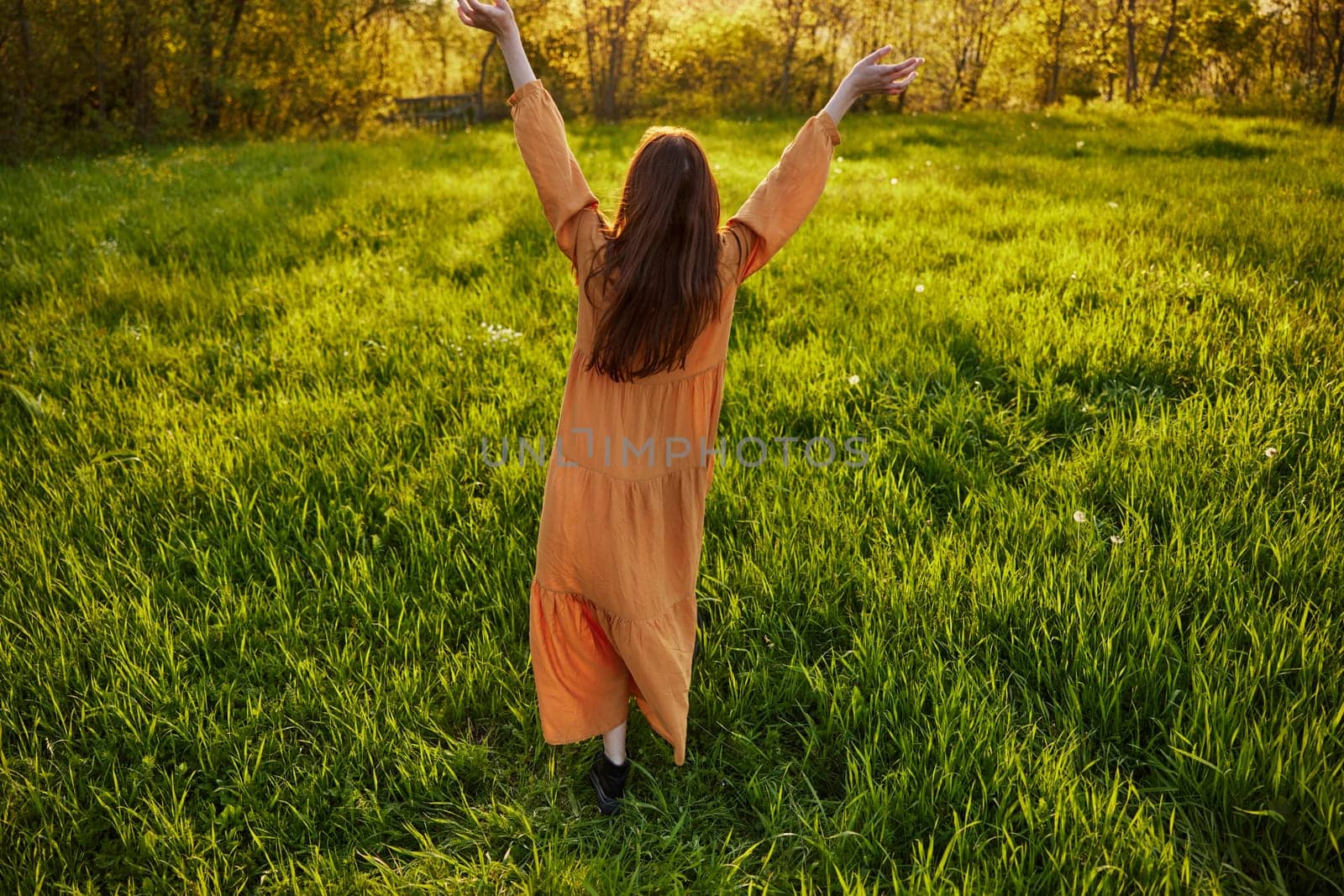 red-haired woman stands in a wide, green field during sunset in a long orange dress enjoying unity with nature and relaxation raising her arms to the sides while standing with her back to the camera by Vichizh