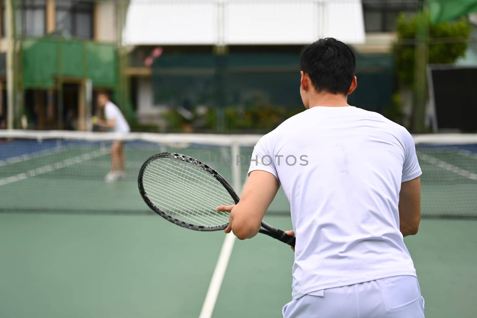 Back view of male tennis player focused in ready position to receive a serve, practicing for competition on a court by prathanchorruangsak