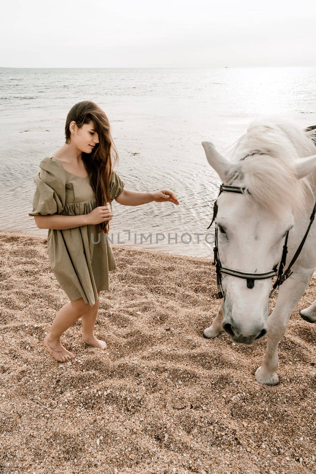 A white horse and a woman in a dress stand on a beach, with the sky and sea creating a picturesque backdrop for the scene. by Matiunina