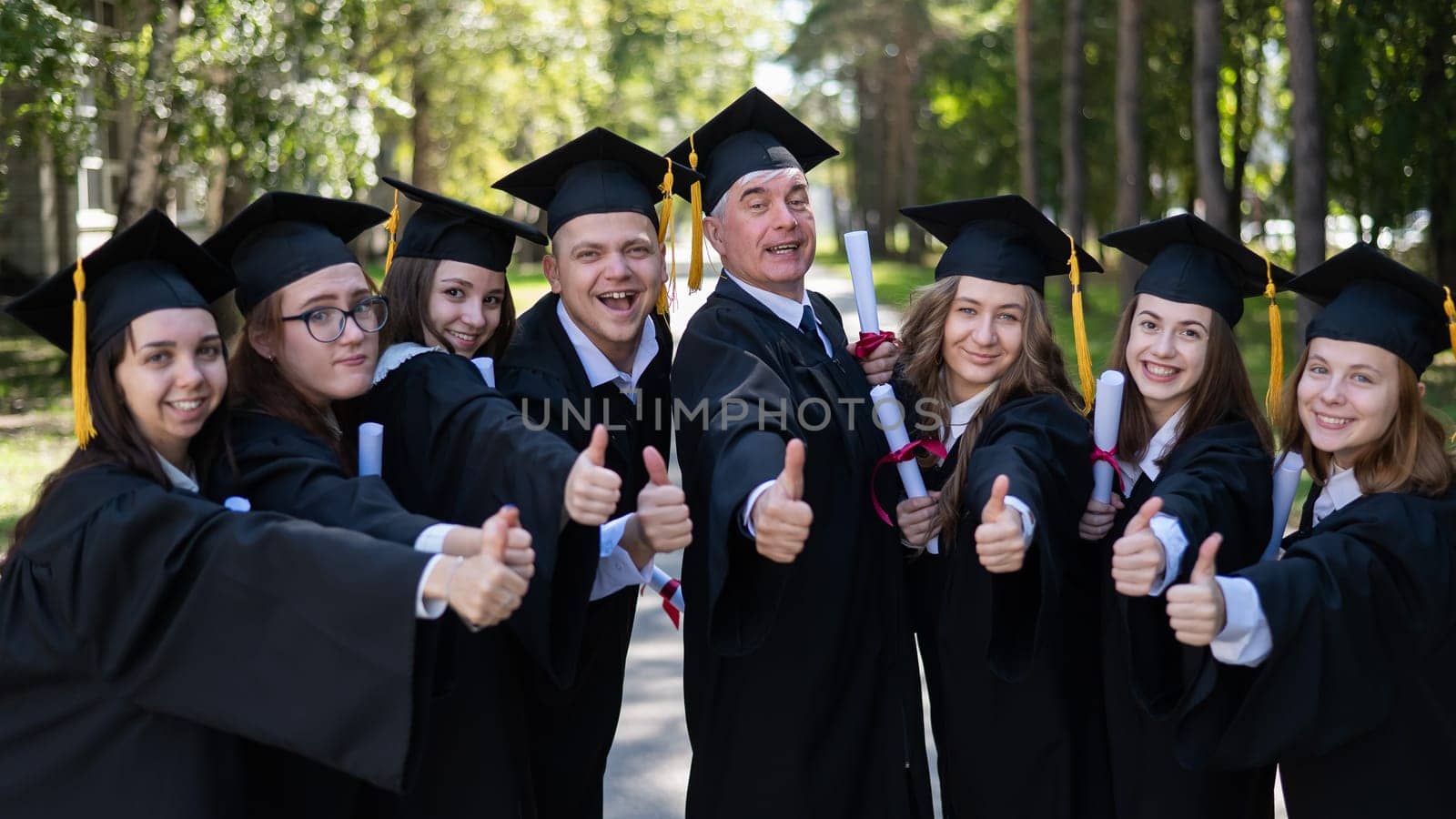 A group of graduates in robes give a thumbs up outdoors. Elderly student. by mrwed54
