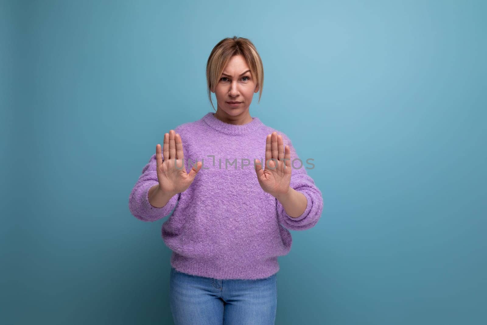 blonde woman in purple hoodie showing stop sign with hands on blue background.