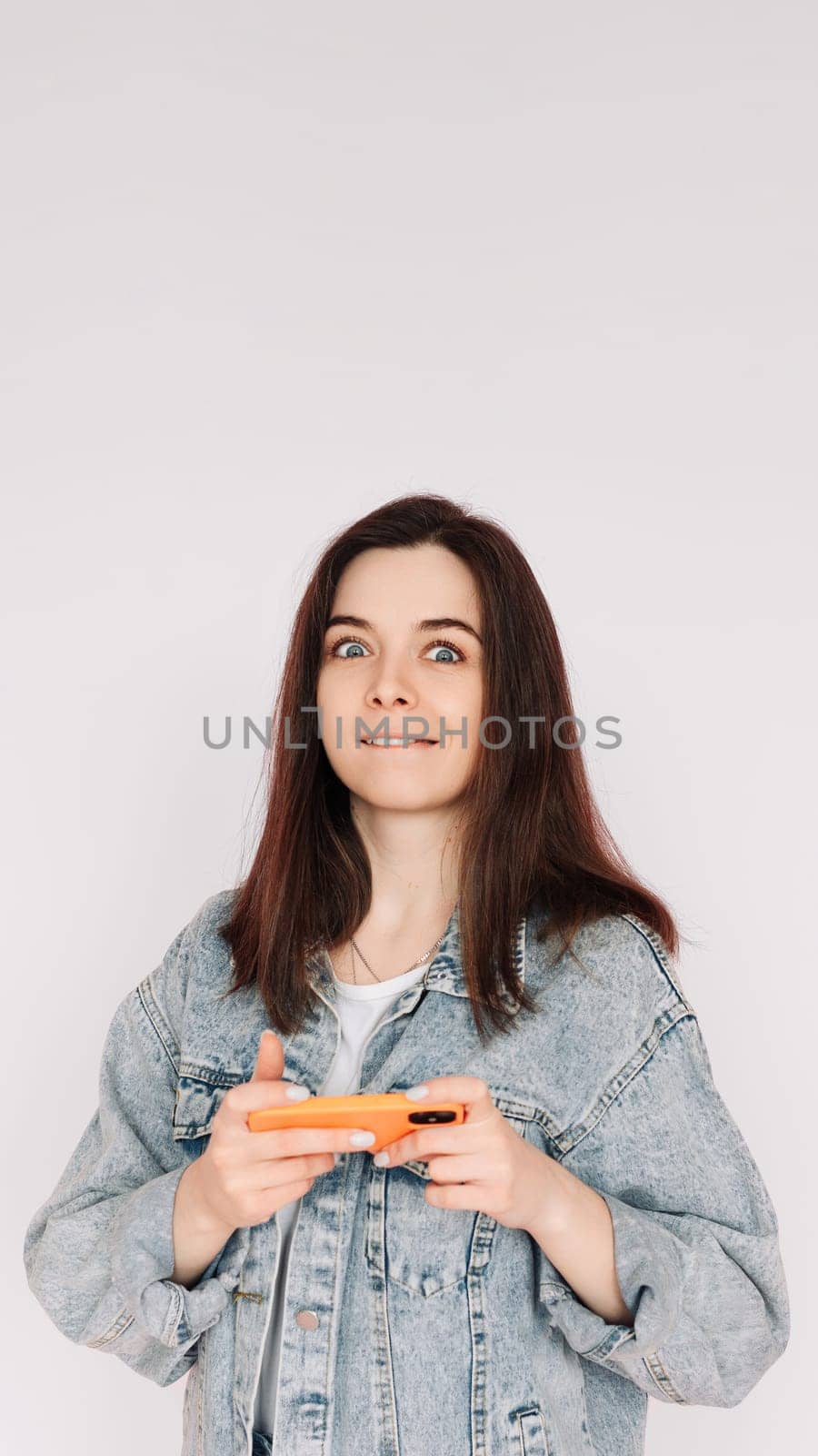 Portrait of a young woman in a casual denim jacket with a smartphone playing a game, isolated against a gray background, conveying indecision and insecurity. Vertical photography