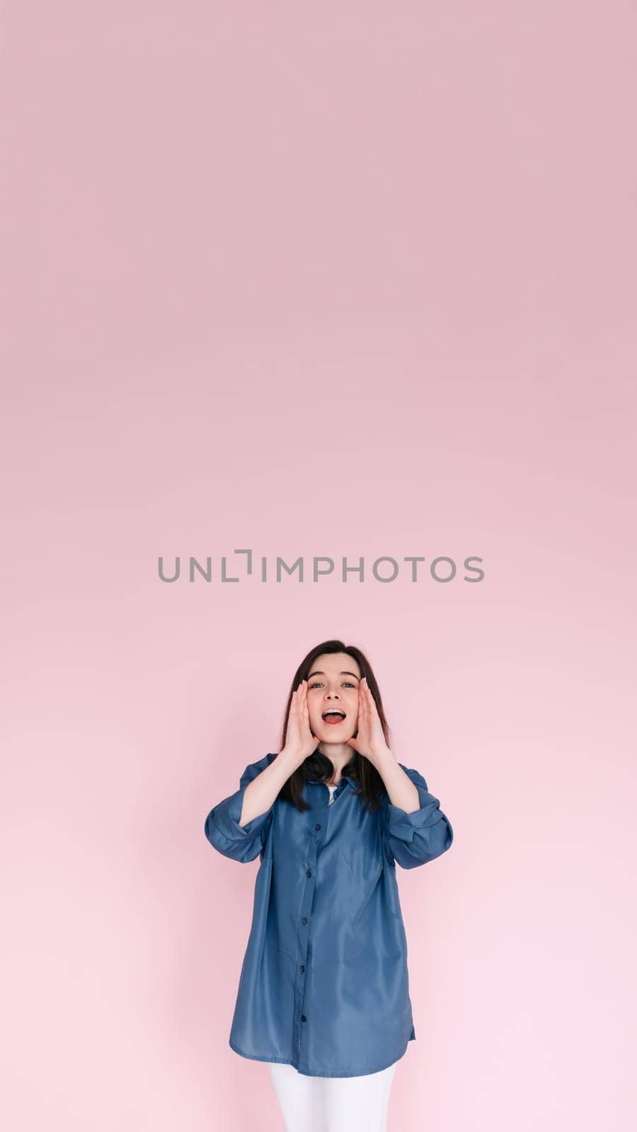 Portrait of young woman with joyous expression, hands near open mouth as if sharing news or telling a funny story, beaming a big toothy smile, isolated on bright pink background, Vertical photography