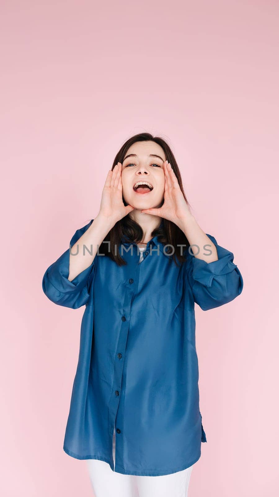 Expressive Joy: Close-Up Portrait of a Cheerful Young Woman's Hands near Open Mouth, Conveying Excitement and Happiness with a Toothy Smile, Isolated on Vibrant Pink Background. Vertical photography.