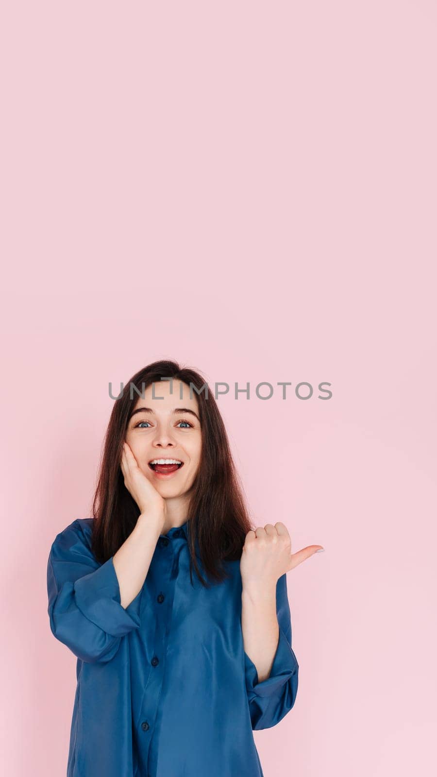 Young woman expressing amazement and joy, touching cheek and pointing finger at empty space, with a big toothy smile on pink isolated background. Vertical photography.