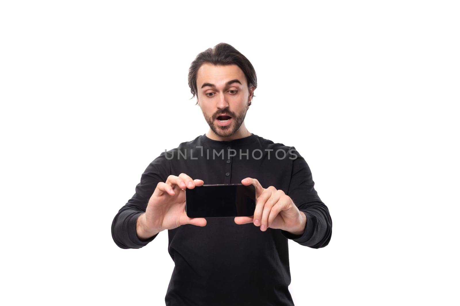 handsome european macho man with black hair and beard with an earring in his ear in surprise shows the screen of a smartphone horizontally standing on an isolated white background with copy space.