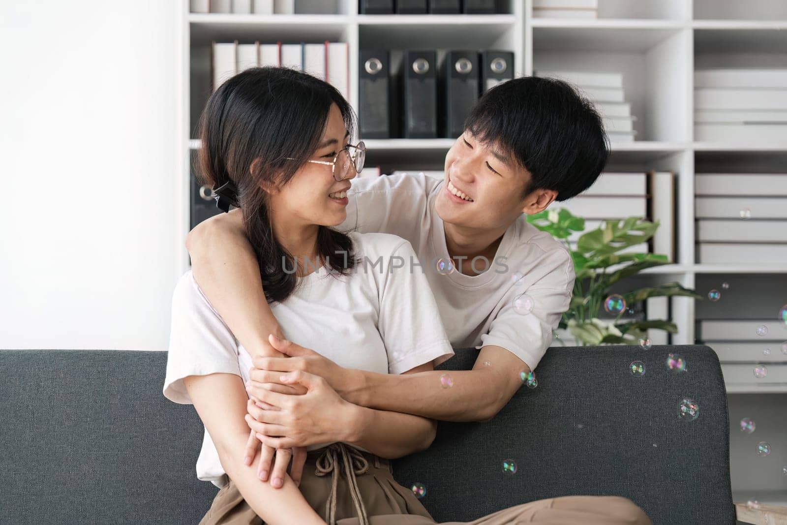 Happy young asian couple relax on cozy sofa at home hugging look at each other, enjoy weekend at home by nateemee