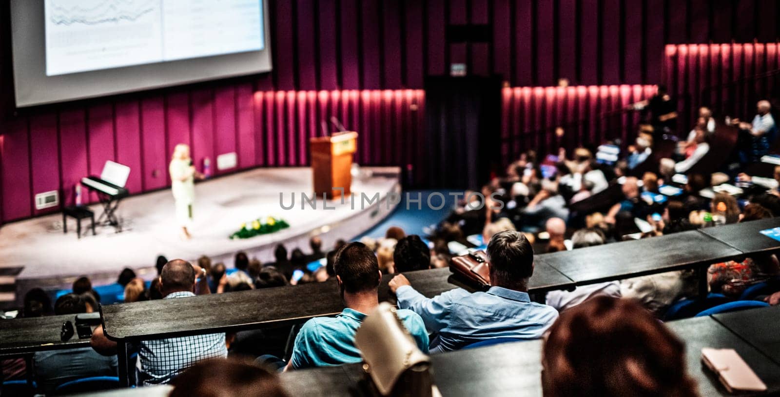 Business and entrepreneurship symposium. Female speaker giving a talk at business meeting. Audience in conference hall. Rear view of unrecognized participant in audience.