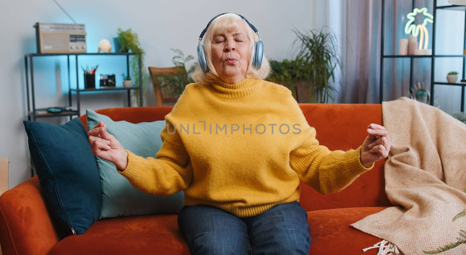 Keep calm down, relax, inner balance. Senior woman breathes deeply with mudra gesture, eyes closed, meditating with concentrated thoughts, peaceful mind at home. Elderly grandmother sitting on couch