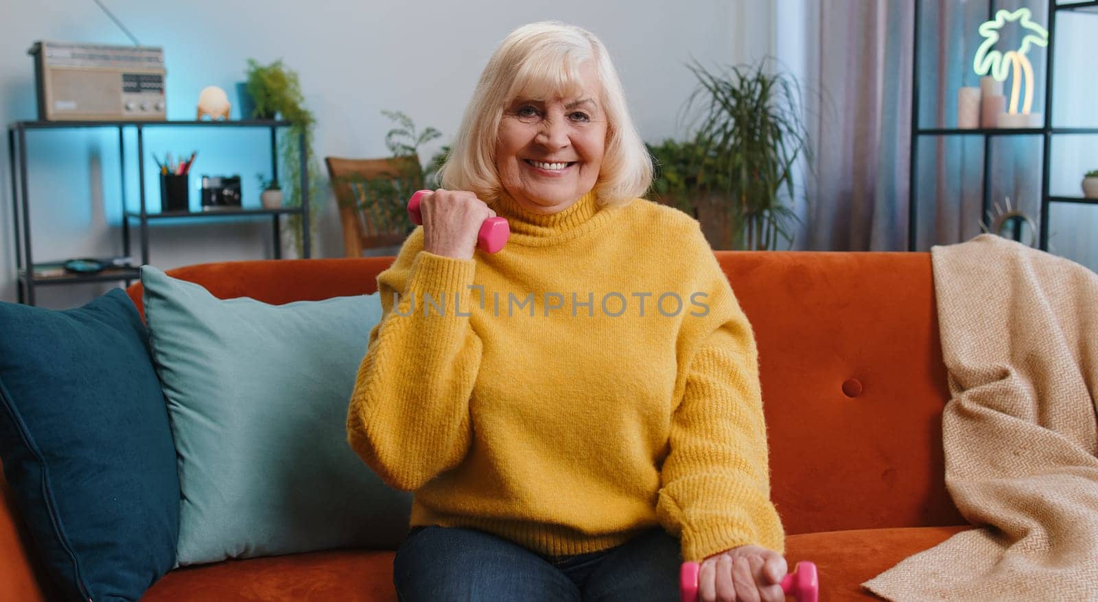 Senior grandmother woman doing dumbbells weight lifting fitness exercises at home apartment indoors. Mature old pensioner workout training strength practicing effort in room on sofa. Sport motivation