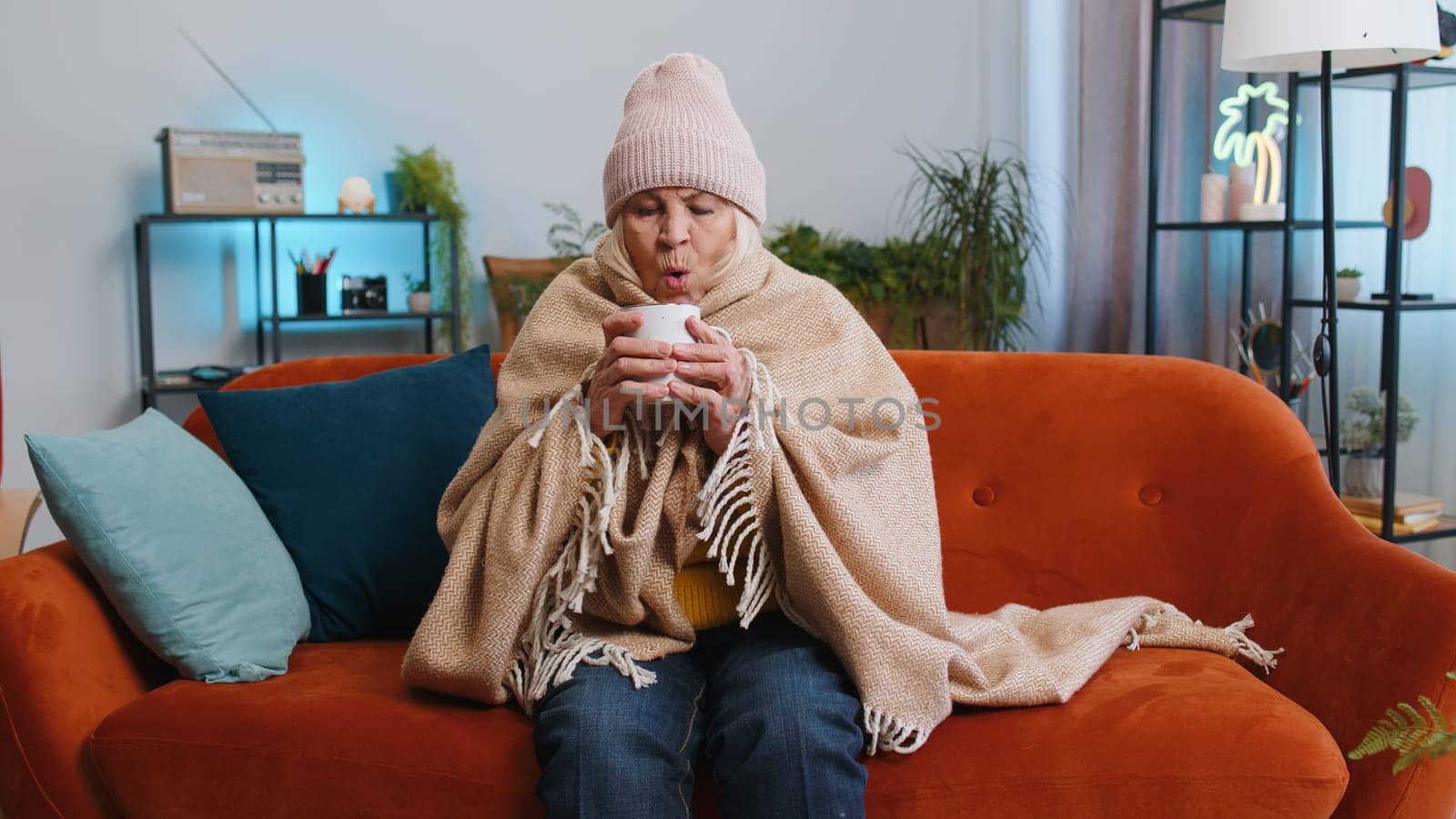 Sick senior grandmother woman in plaid shivering from cold on sofa drinking hot tea in unheated apartment without heating due debt. Unhealthy ill elderly pensioner feeling discomfort try to warming up