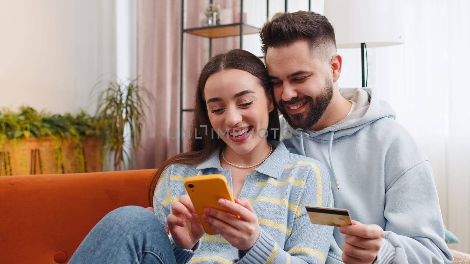 Family married couple man woman using credit bank card and smartphone while transferring money, purchases online shopping, order food delivery at home. Husband wife together on couch in living room