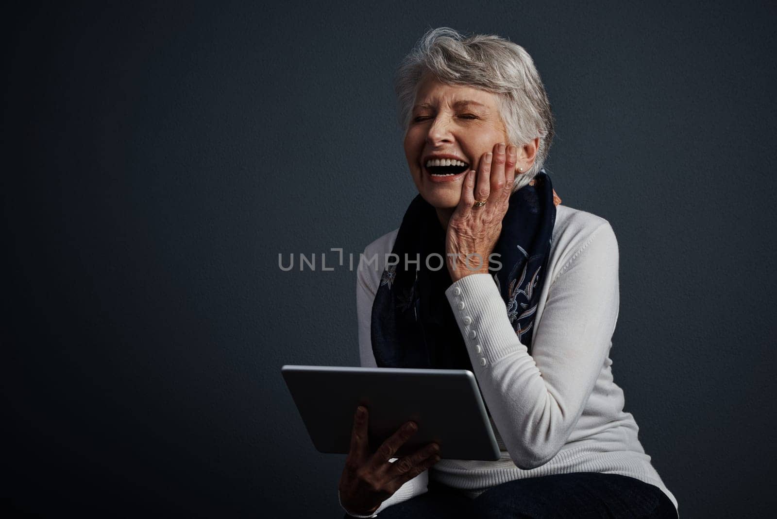 This thing cracks me up. Studio shot of an cheerful elderly woman sitting and using a tablet. by YuriArcurs