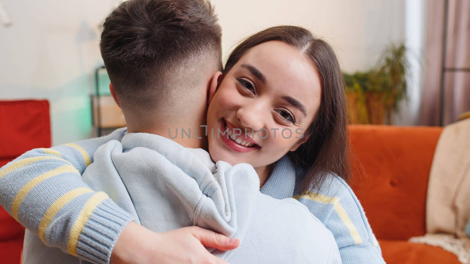 Portrait of woman embracing, hugging man, looking at camera over his shoulder at home. Young authentic lovely family marriage couple. Partnership, trust and support. Husband and wife together in room