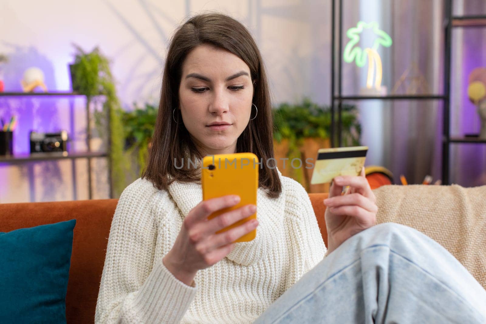 Young woman using credit bank card and smartphone while transferring money, purchases online shopping cashless, order food delivery at home apartment indoors. Happy girl sitting in room on couch