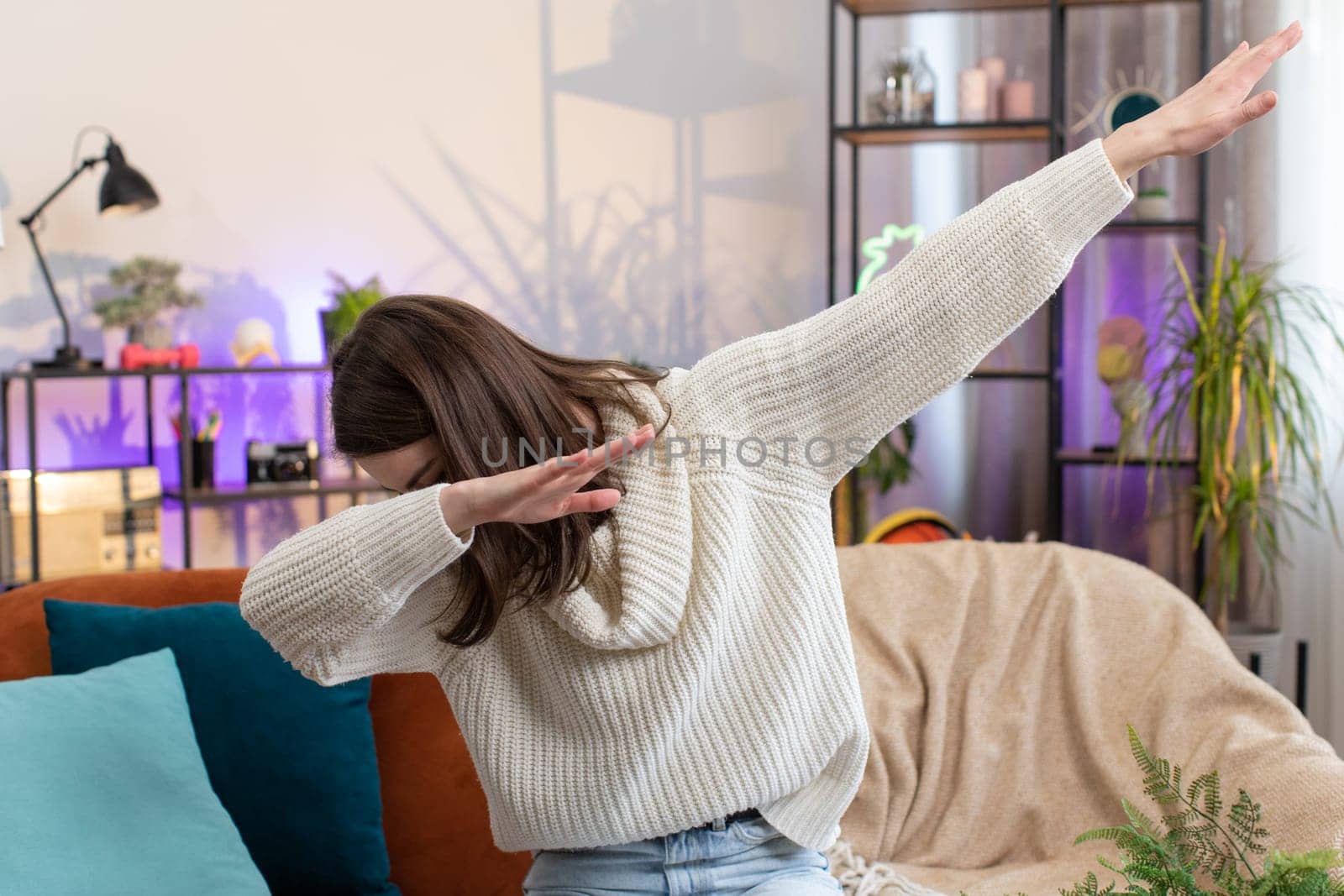 Portrait of trendy cheerful positive young woman having fun dancing and moving to rhythm, dabbing raising hands, making dub dance social media gesture at home room apartment. Pretty girl sits on couch