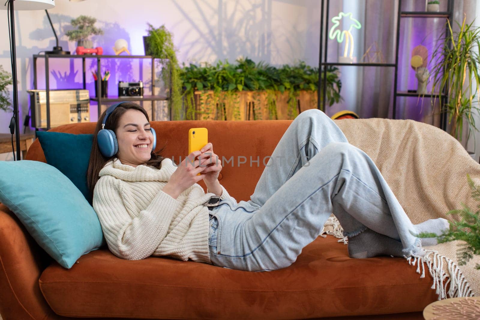 Portrait of happy overjoyed woman in wireless headphones relaxing lying on couch at home apartment choosing listening favorite energetic disco dancing music. People weekend daytime leisure activities