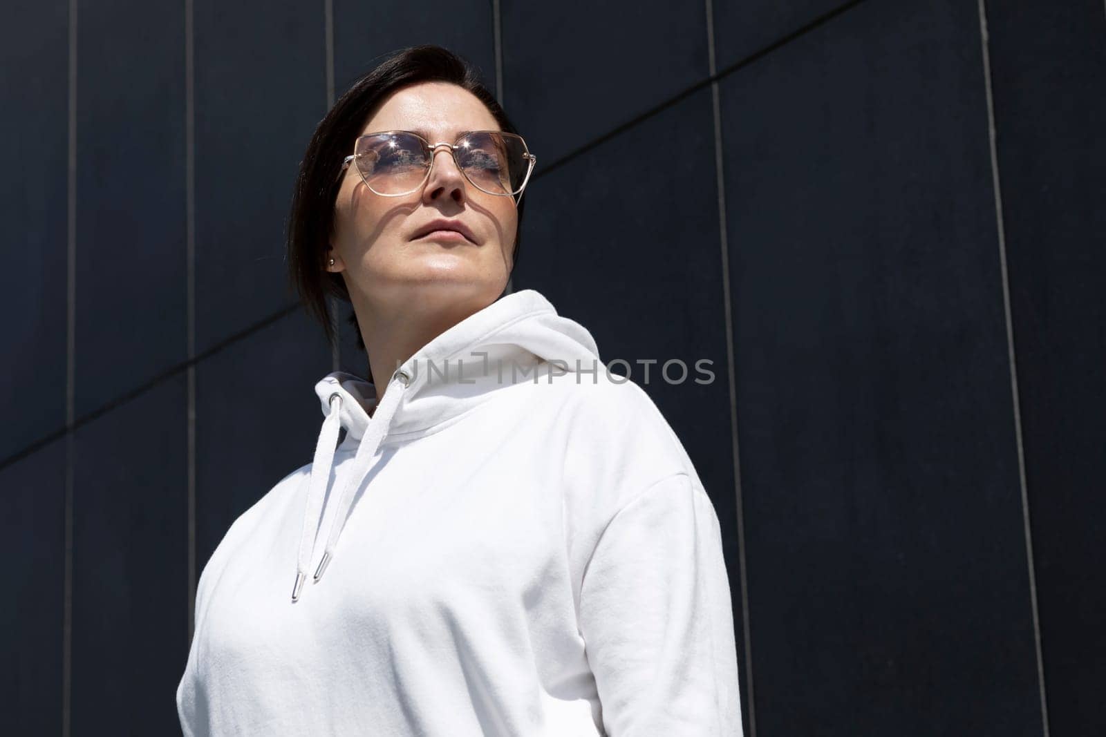 Portrait Pretty Mature Brunette Woman in White Hoodie and Sunglasses Looks Away, Dark Background Wall. Sportive Confident 40 Yo Beautiful Caucasian Woman in City. Horizontal Plane . High quality photo