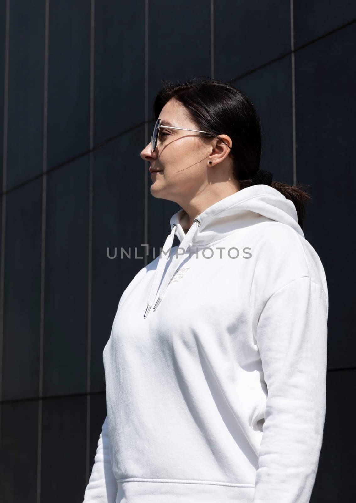 Portrait Pretty Mature Brunette Woman in White Hoodie and Sunglasses Looks Away, Dark Background Wall. Sportive Confident 40 Yo Beautiful Caucasian Woman in City. Vertical Plane . High quality photo