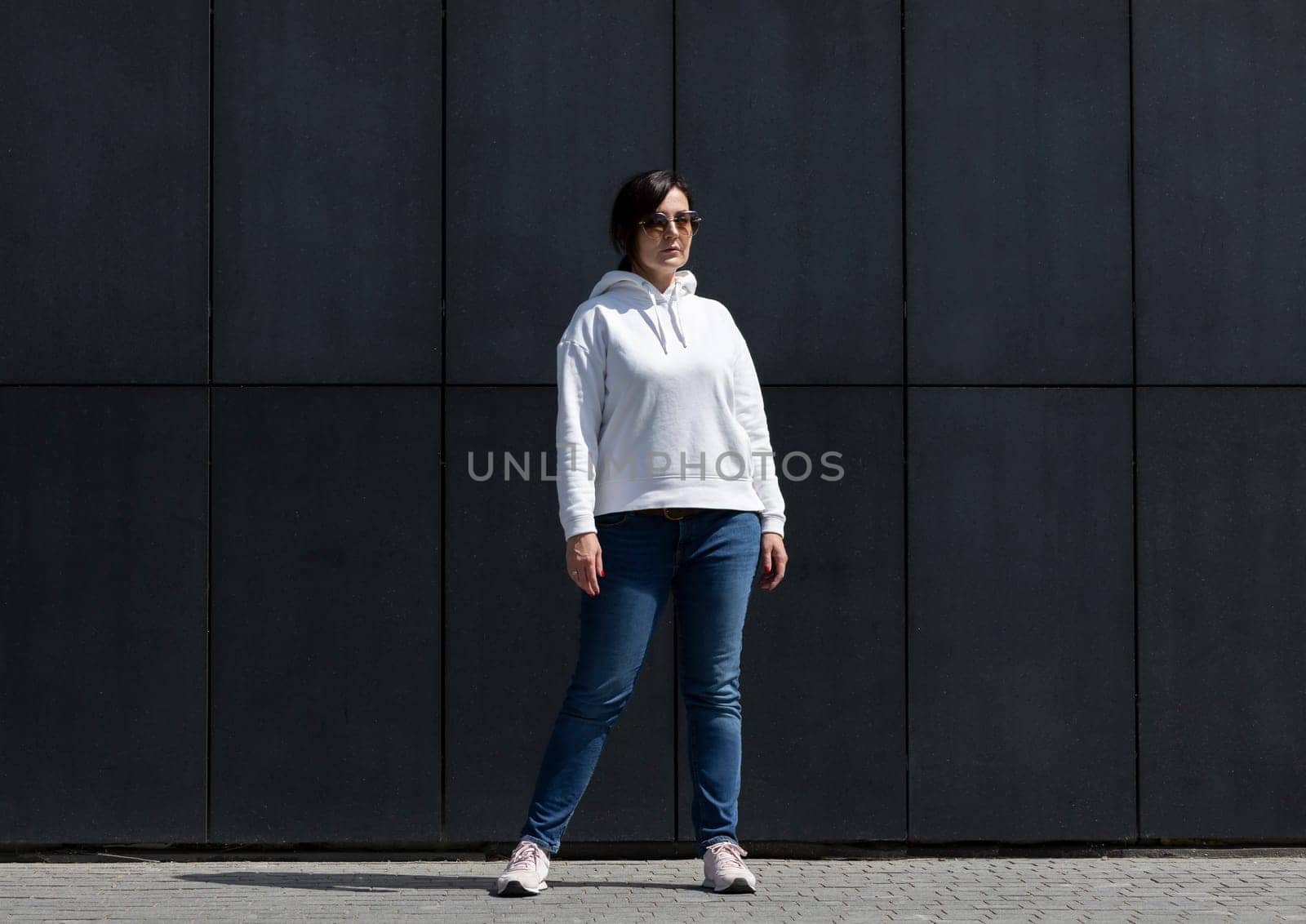 Full Length Pretty Mature Brunette Woman in White Hoodie and Sunglasses Looks Away, Dark Background Wall. Sportive Confident 40 Yo Beautiful Caucasian Woman in City. Horizontal,High quality photo