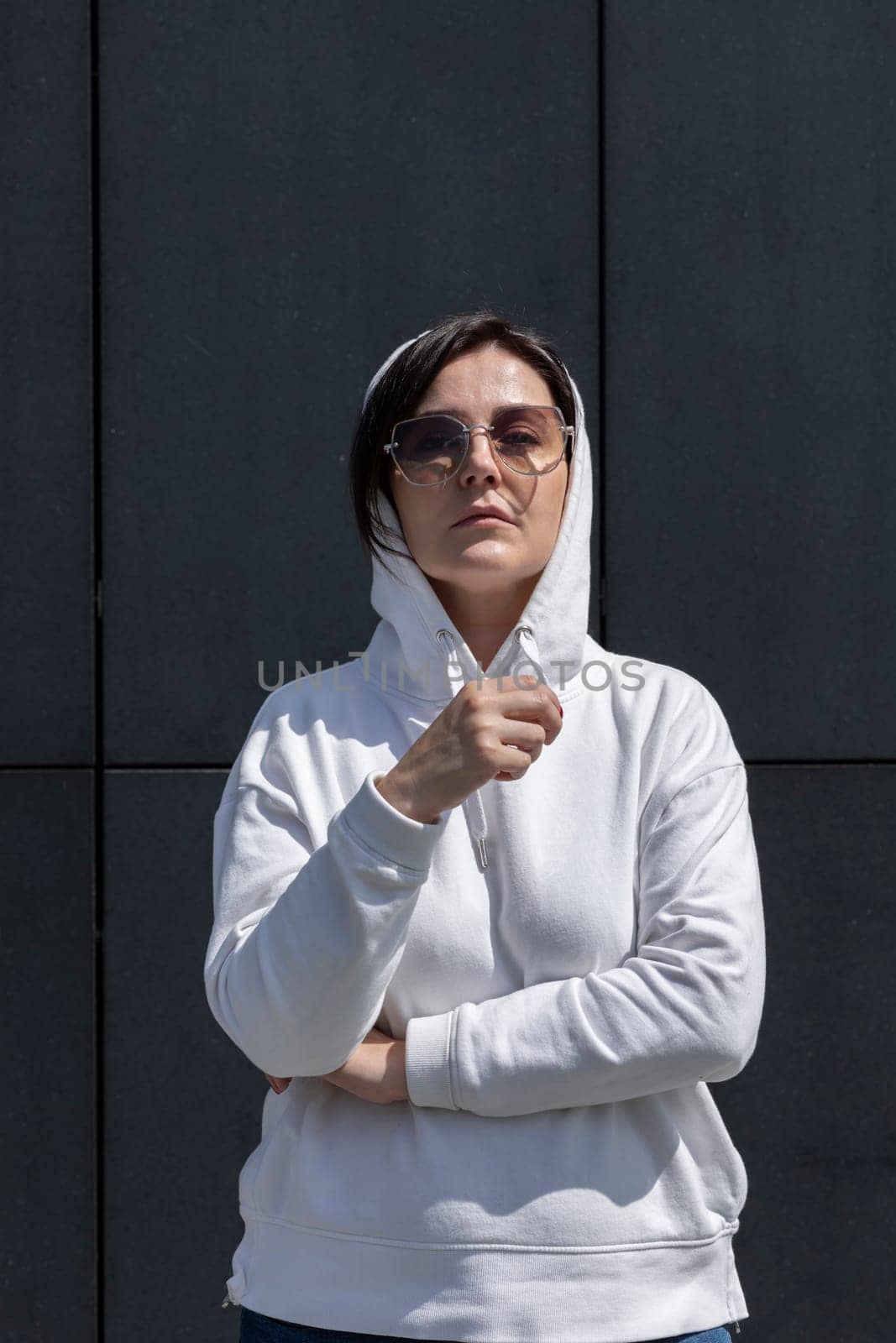 Portrait Mature Brunette Woman With Hood On Head, Wears White Hoodie and Sunglasses Looks Straight, Dark Background Wall. Sportive Confident 40 Yo Beautiful Caucasian Woman Outdoor. Vertical Plane.