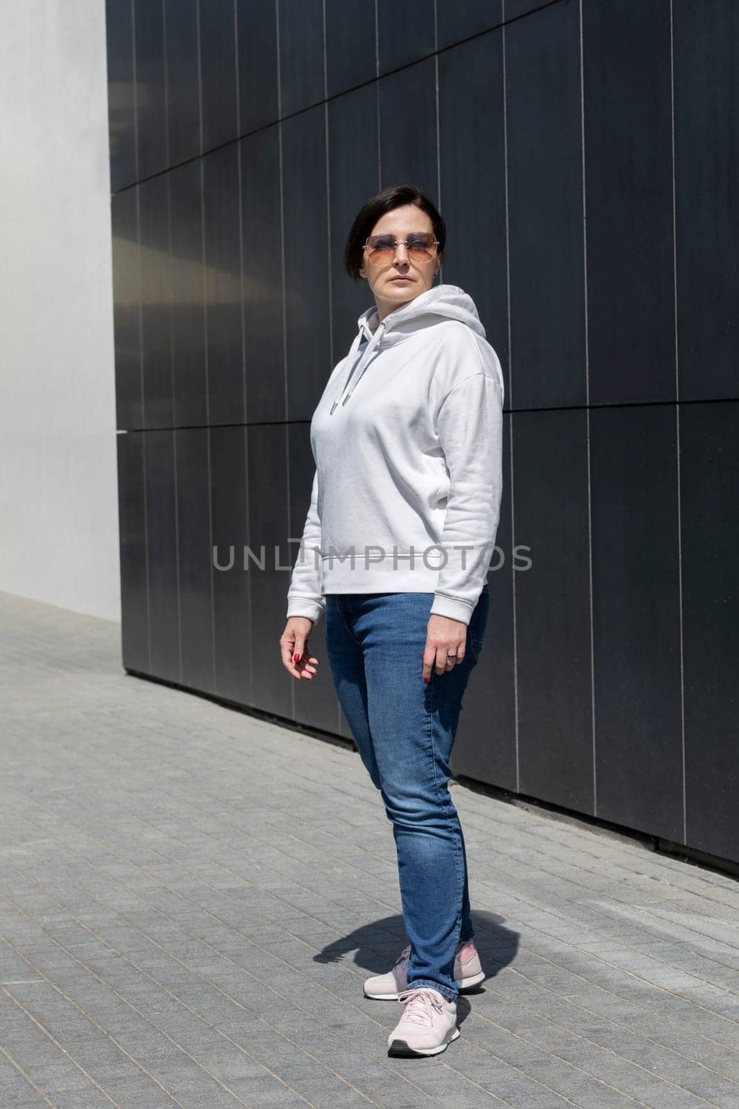 Full Length Pretty Mature Brunette Woman in White Hoodie, Jeans and Sunglasses Looks Away, Dark Background Wall. Sportive Confident 40 Yo Beautiful Caucasian Woman in Urban Area Vertical Plane.