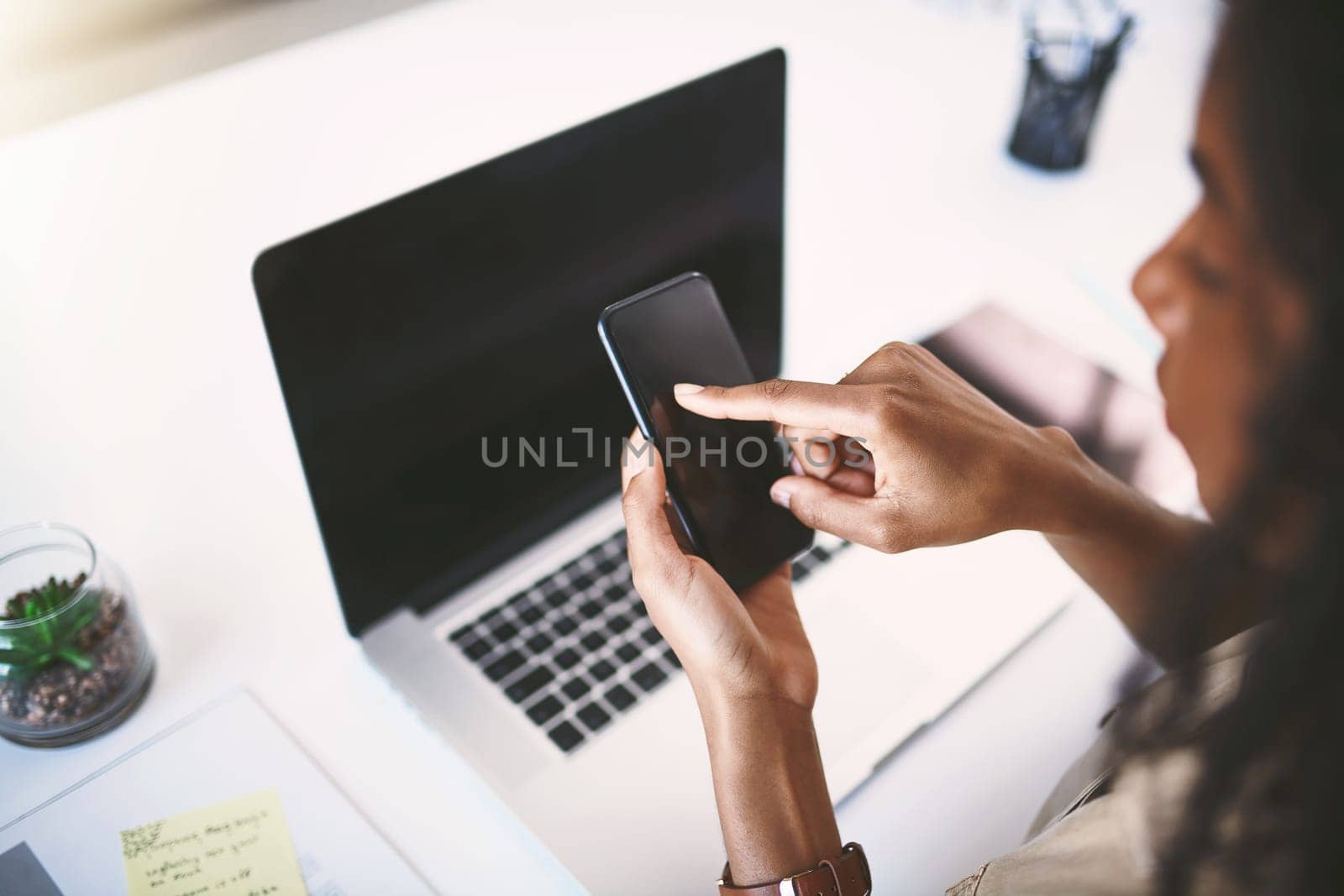 Making success happen with smart apps. a young businesswoman using a mobile phone and laptop at her desk in a modern office. by YuriArcurs