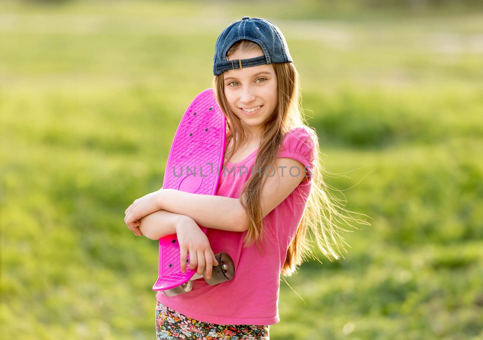 Attractive teenage girl with a cap on holding her bright pink skating board, smiling