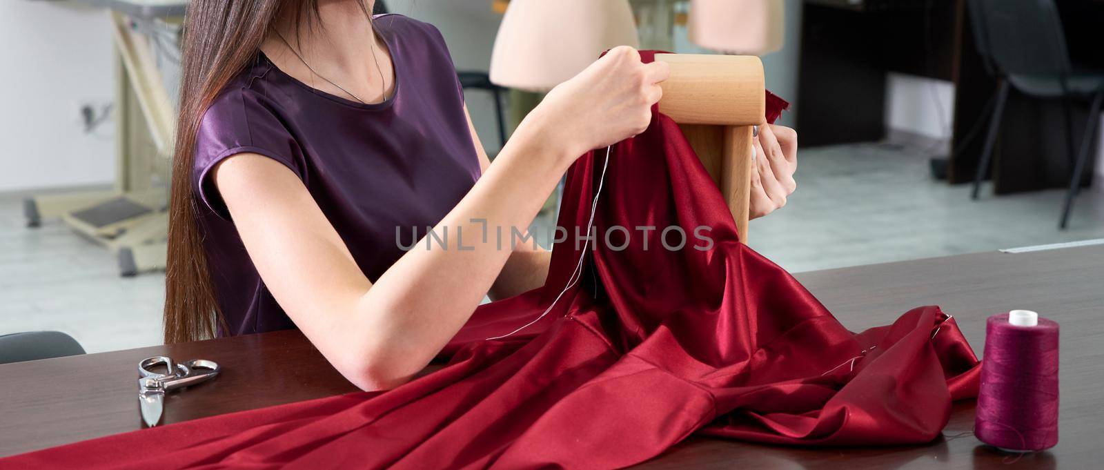 Beautiful young dressmaker in workroom sewing red dress in tailor studio or atelier shop by Mariakray