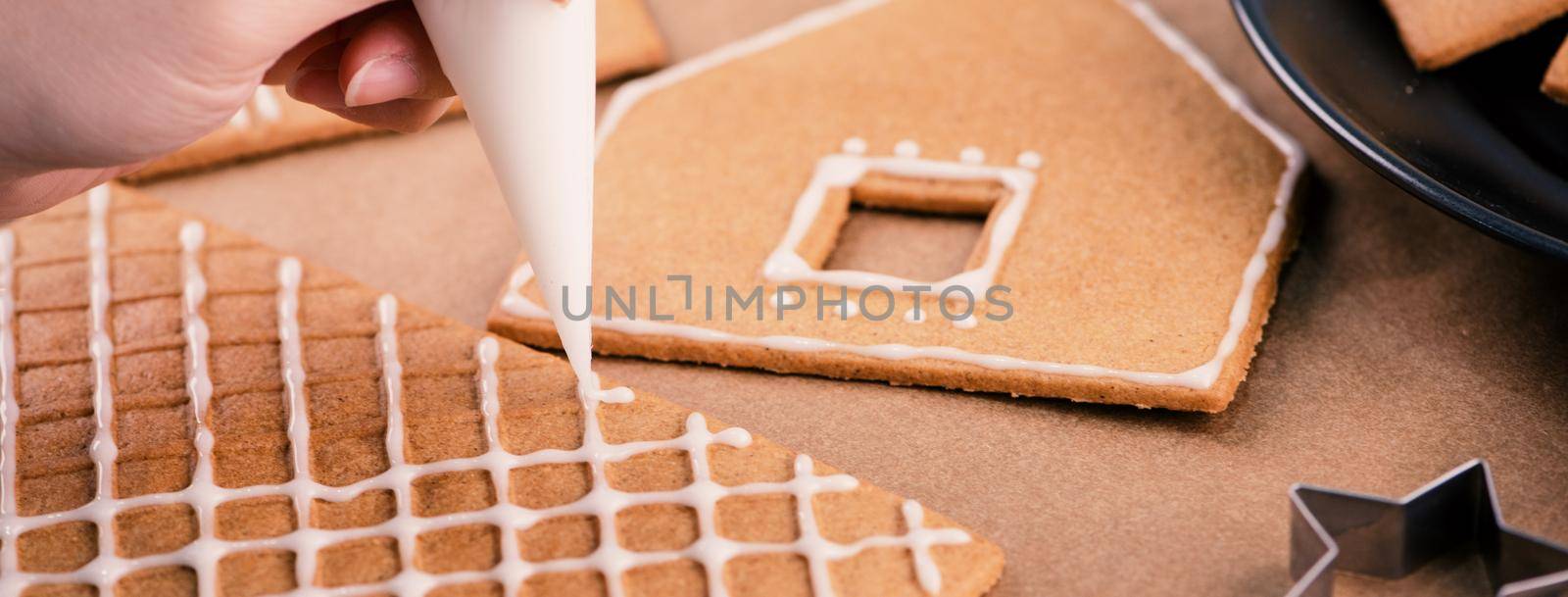 Woman is decorating gingerbread cookies house with white frosting icing cream topping on wooden table background, baking paper in kitchen, close up, macro.