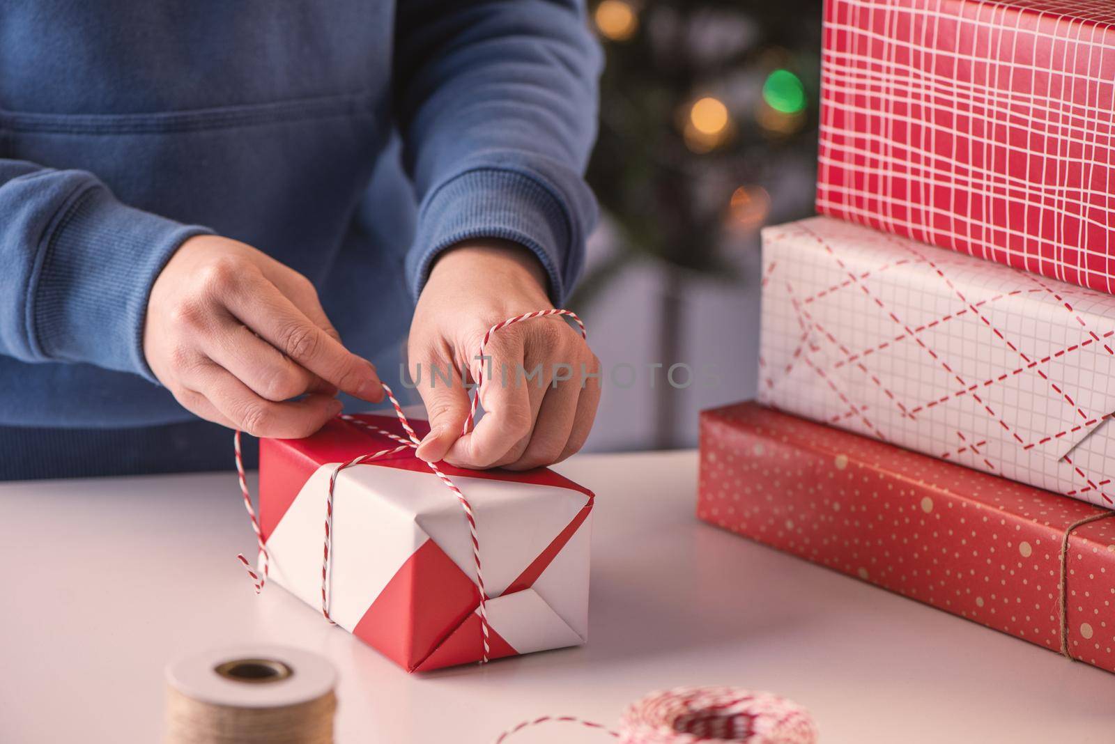 A young woman in blue is wrapping, packaging Christmas gift for kids children by beautiful, red, white wrapper with glittering light, lifestyle.