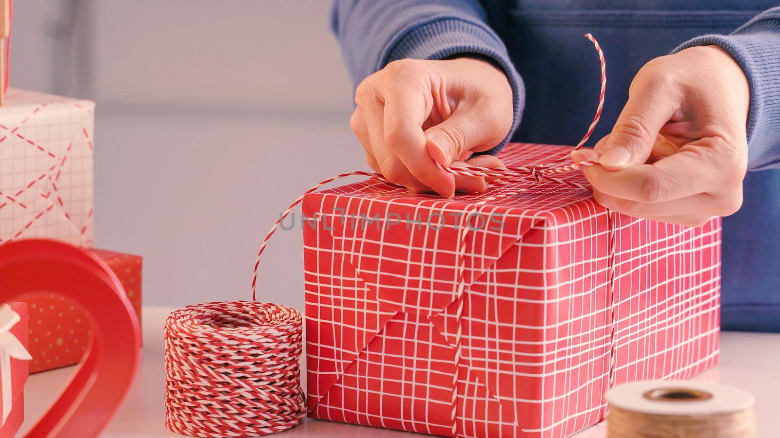 A young woman in blue is wrapping, packaging Christmas gift for kids children by beautiful, red, white wrapper with glittering light, lifestyle.