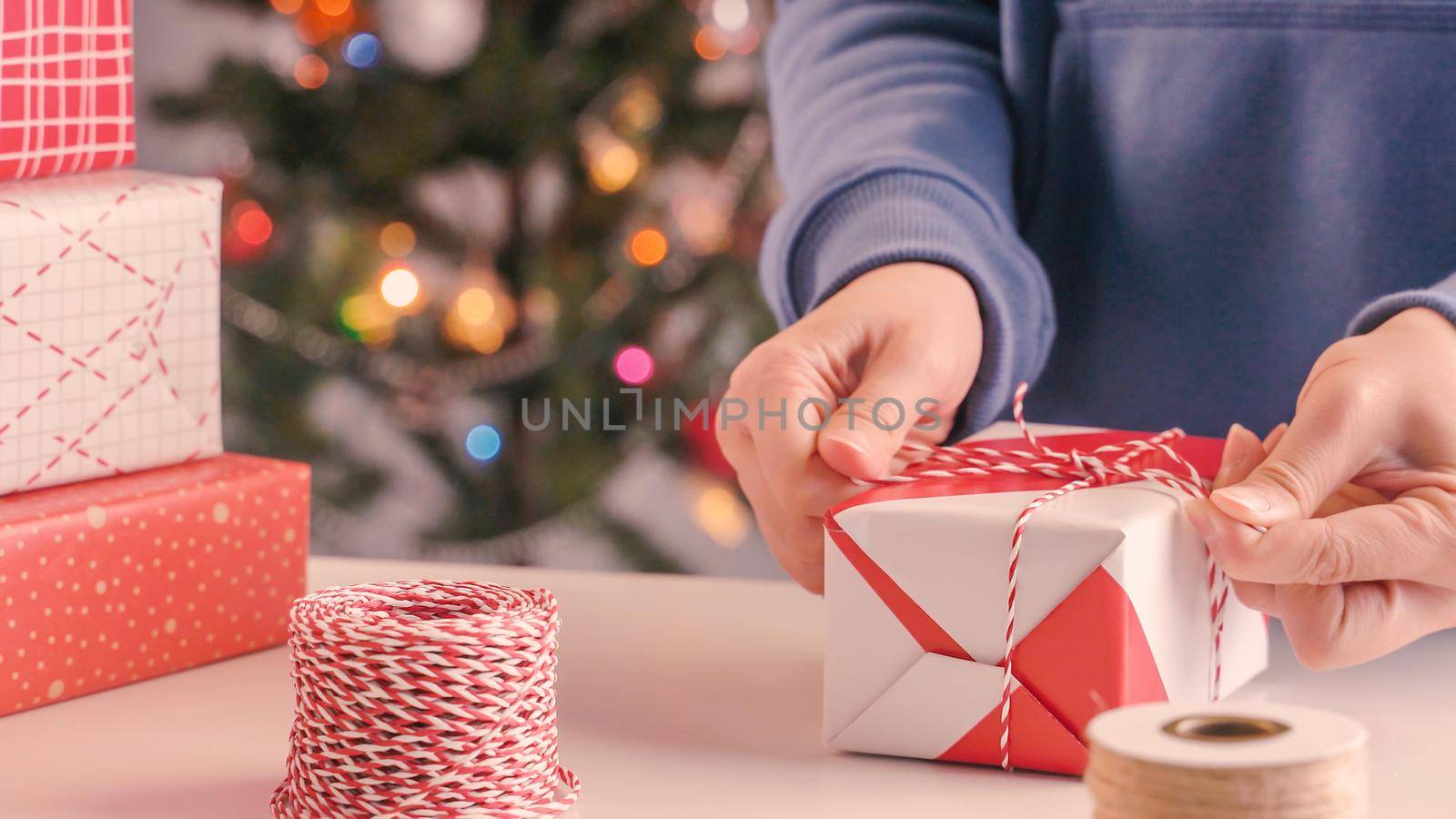 A young woman in blue is wrapping, packaging Christmas gift for kids children by beautiful, red, white wrapper with glittering light, lifestyle.