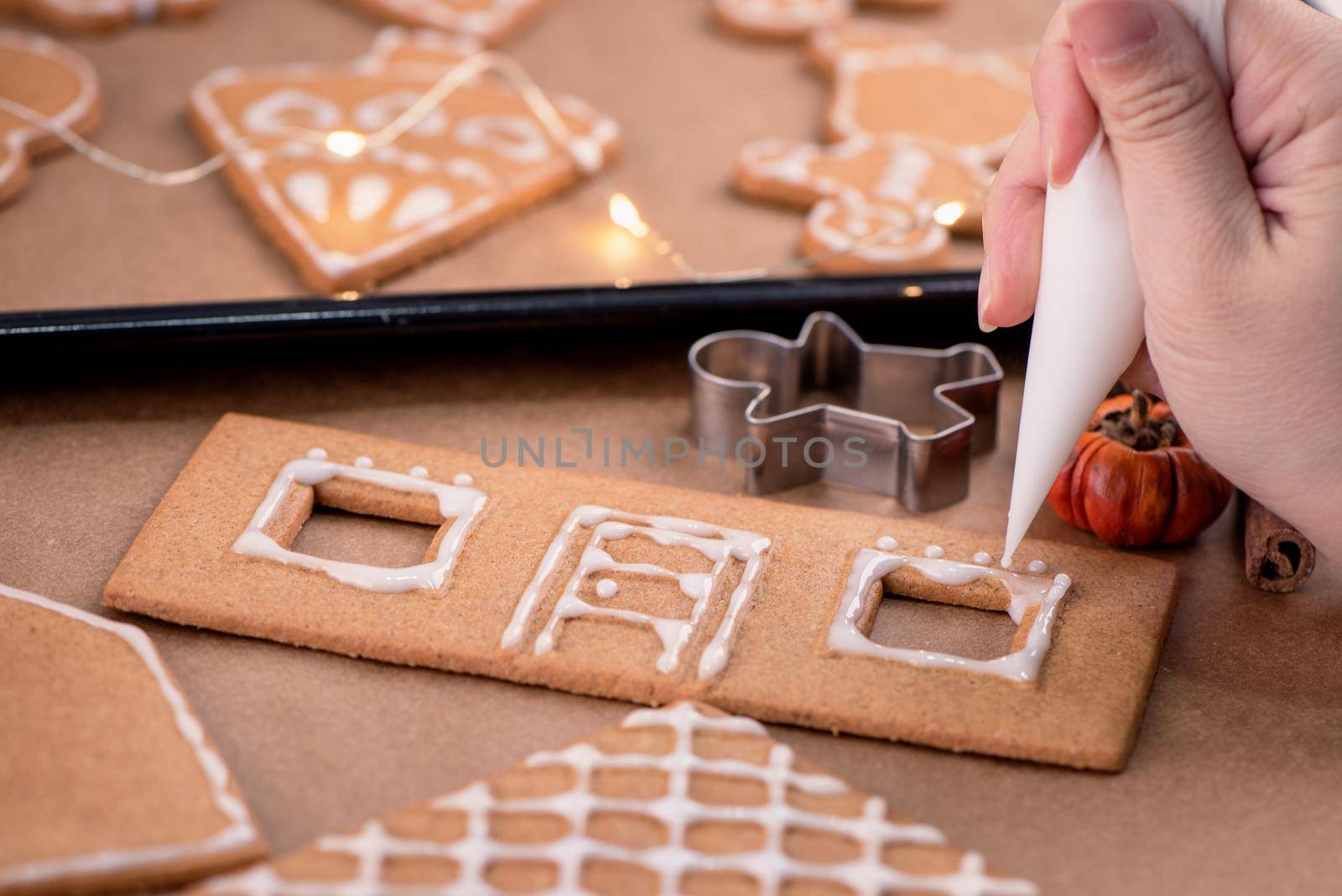 Woman is decorating gingerbread cookies house with white frosting icing cream topping on wooden table background, baking paper in kitchen, close up, macro.