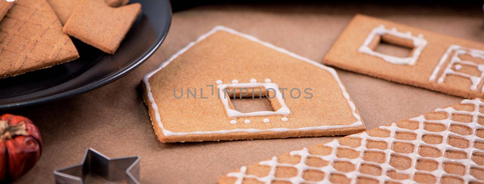 Woman is decorating gingerbread cookies house with white frosting icing cream topping on wooden table background, baking paper in kitchen, close up, macro.