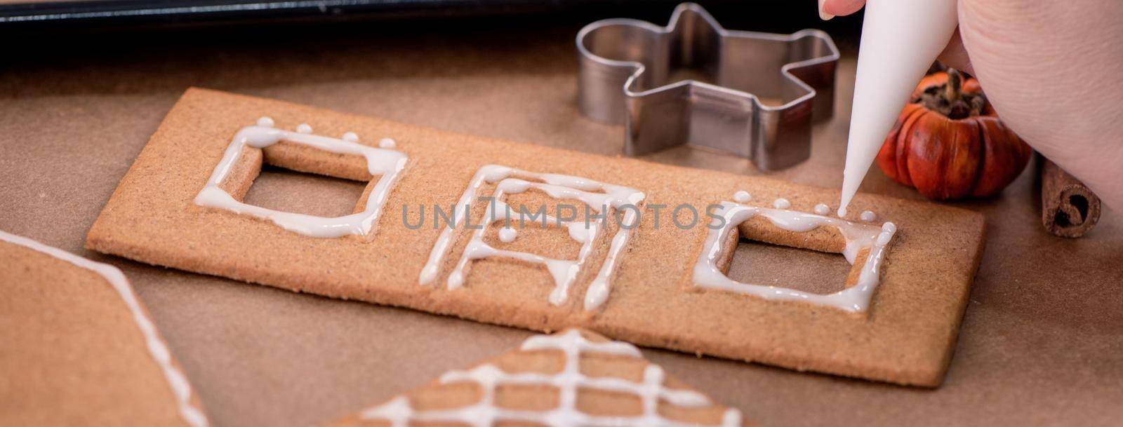 Woman is decorating gingerbread cookies house with white frosting icing cream topping on wooden table background, baking paper in kitchen, close up, macro. by ROMIXIMAGE