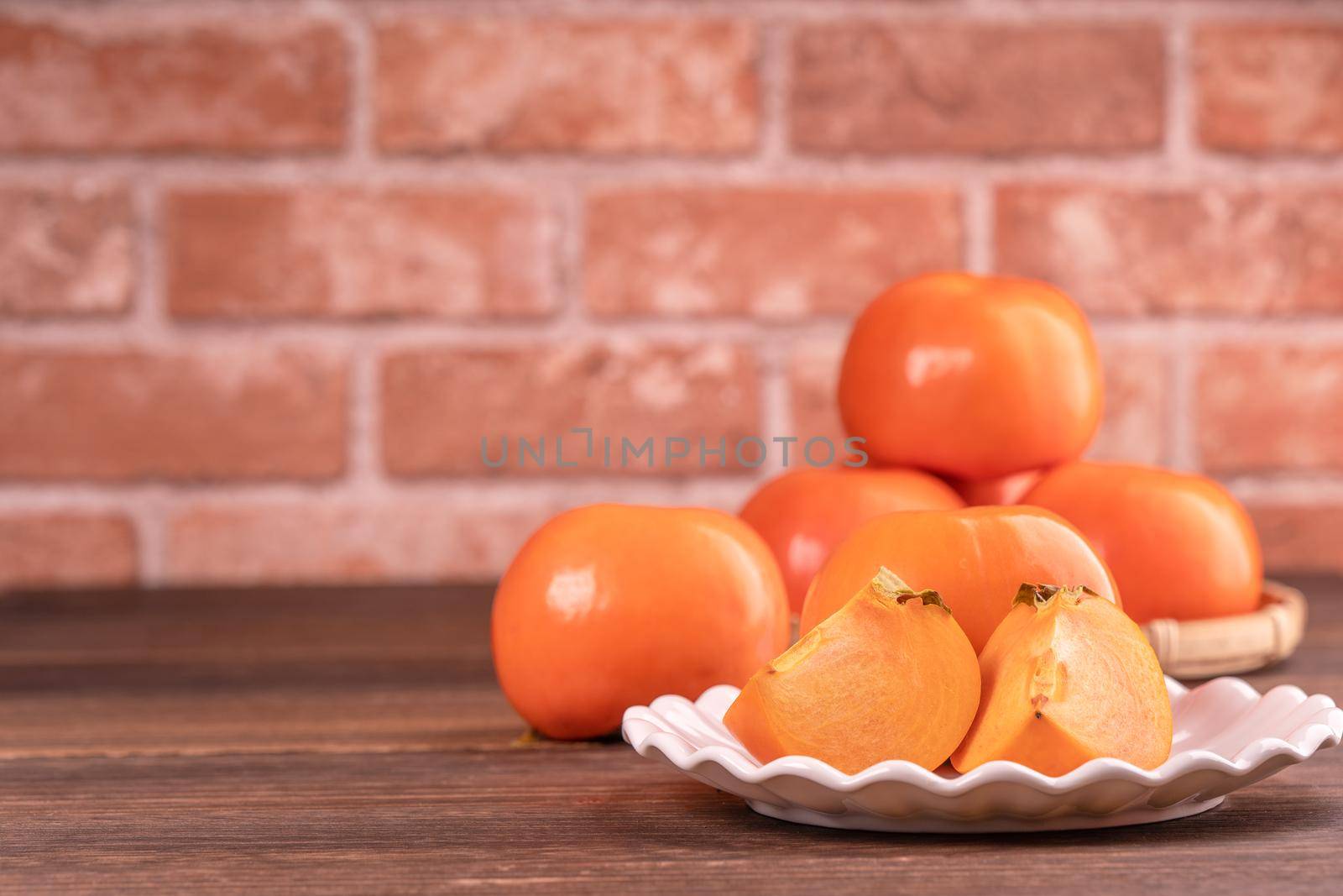 Sliced sweet persimmon kaki in a bamboo sieve basket on dark wooden table with red brick wall background, Chinese lunar new year fruit design concept, close up.
