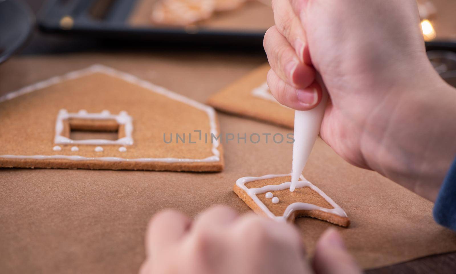 Woman is decorating gingerbread cookies house with white frosting icing cream topping on wooden table background, baking paper in kitchen, close up, macro.