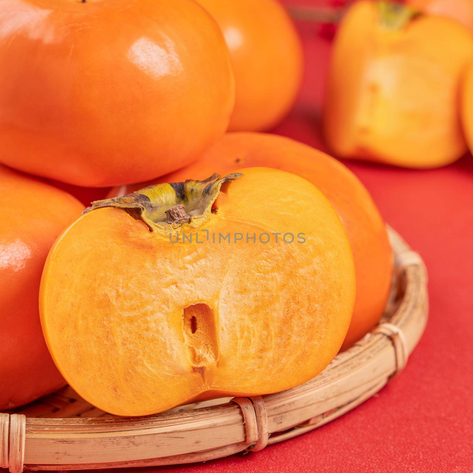 Fresh beautiful sliced sweet persimmon kaki isolated on red table background and bamboo sieve, Chinese lunar new year design concept, close up. by ROMIXIMAGE